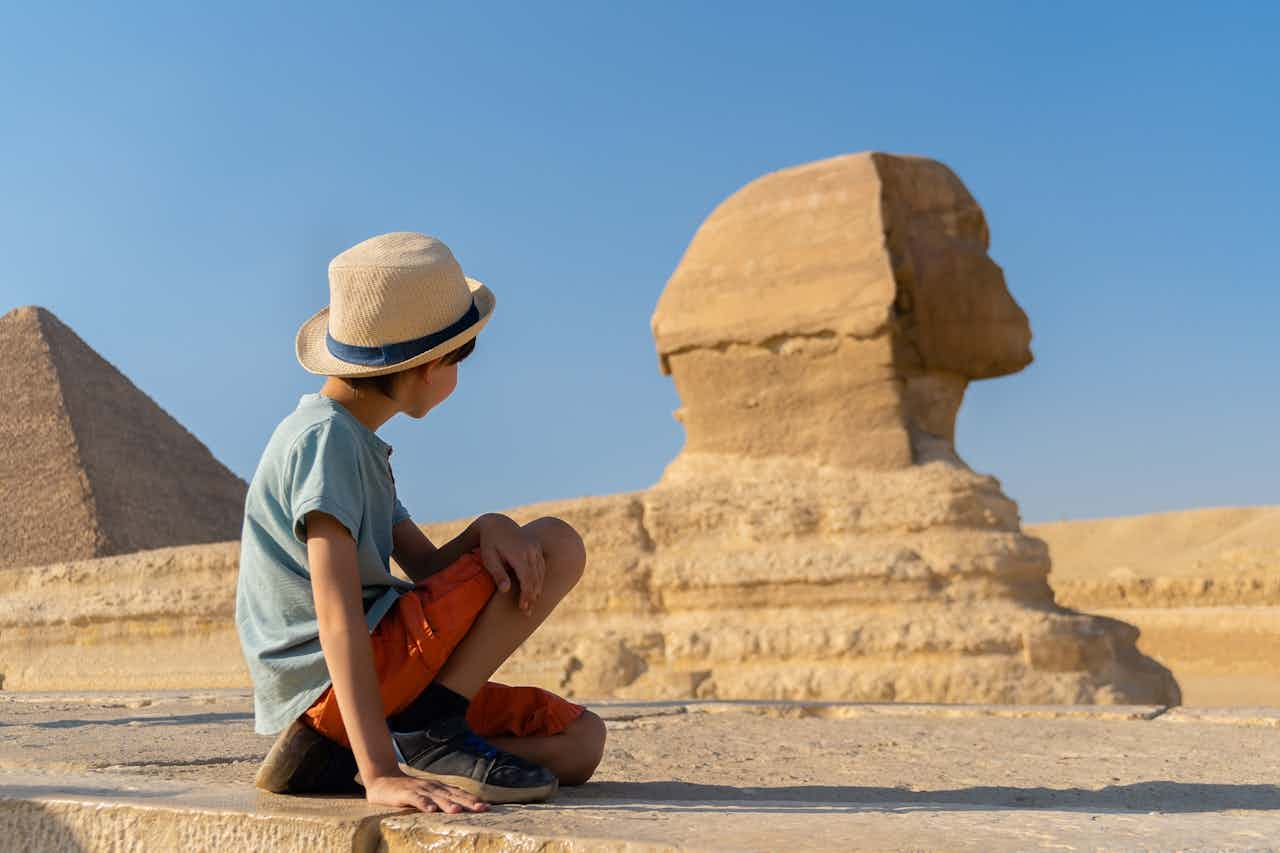 Magischer Moment in Ägypten: Ein Junge mit Strohhut sitzt vor der majestätischen Sphinx von Gizeh und bestaunt die jahrtausendealte Geschichte. Im Hintergrund ragt eine der legendären Pyramiden in den strahlend blauen Himmel. Ein unvergessliches Reiseerlebnis für Kulturliebhaber, die das Geheimnis und den Luxus des alten Ägyptens hautnah erleben möchten.