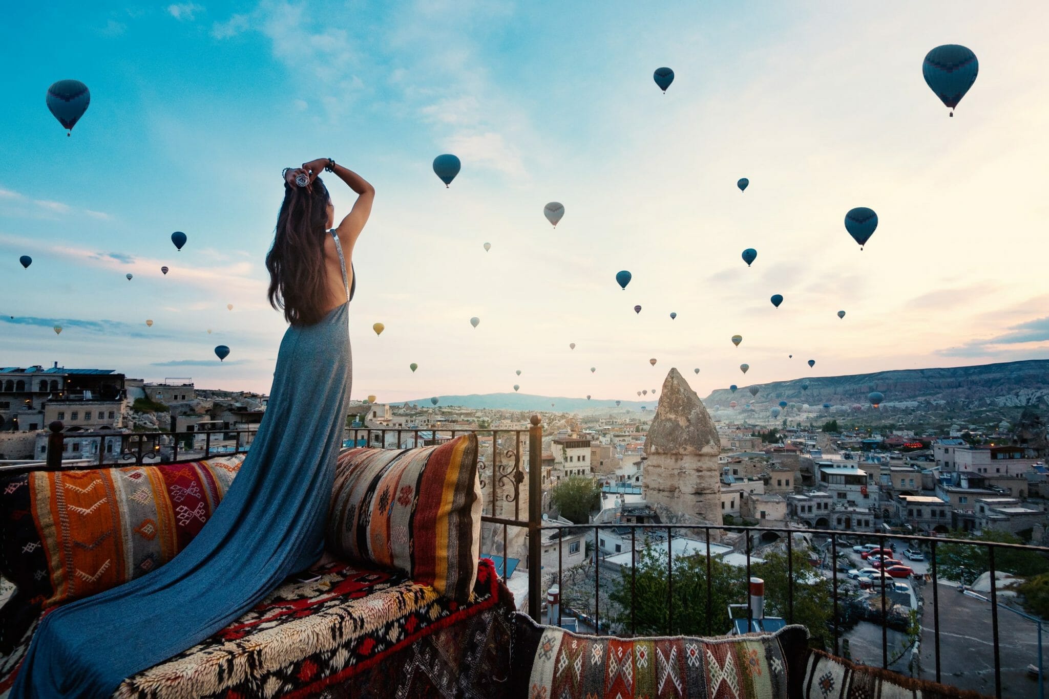 Spektakuläre Heißluftballonfahrt in Kappadokien bei Sonnenaufgang. Eine junge, wunderschöne Frau in einem eleganten, langen Kleid steht inmitten der atemberaubenden Landschaft und blickt auf die schwebenden Ballons. Ein unvergessliches Bucket-List-Erlebnis für Luxusreisende und Abenteurer, die die Magie von Kappadokien hautnah erleben möchten