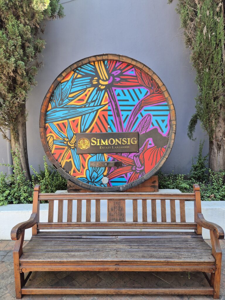 Das Simonsig Wine Estate in Südafrika vereint traditionelle Weinherstellung mit kreativem Design und modernem Lifestyle. Dieses kunstvoll gestaltete Weinfass ist ein Symbol für die Innovationskraft des renommierten Weinguts – ein Ort, an dem Exzellenz und Kreativität miteinander verschmelzen. Der kräftige Farbmix aus Blau-, Lila- und Orangetönen verleiht der rustikalen Szenerie einen modernen Touch und spiegelt die lebendige Weinkultur Südafrikas wider. Simonsig ist bekannt für seine erstklassigen Weine und gilt als Geburtsstätte des südafrikanischen Methode Cap Classique (MCC) Schaumweins. Hier, im Herzen des Stellenbosch-Weinlands, erleben Besucher unvergessliche Weinproben, malerische Weinberge und luxuriöse Genussmomente. Wer auf der Suche nach einem exklusiven Wein-Erlebnis ist, kann mit MHS Reisen eine maßgeschneiderte Südafrika-Tour planen – inklusive der besten Weingüter, spektakulärer Landschaften und unvergesslicher Genussmomente.